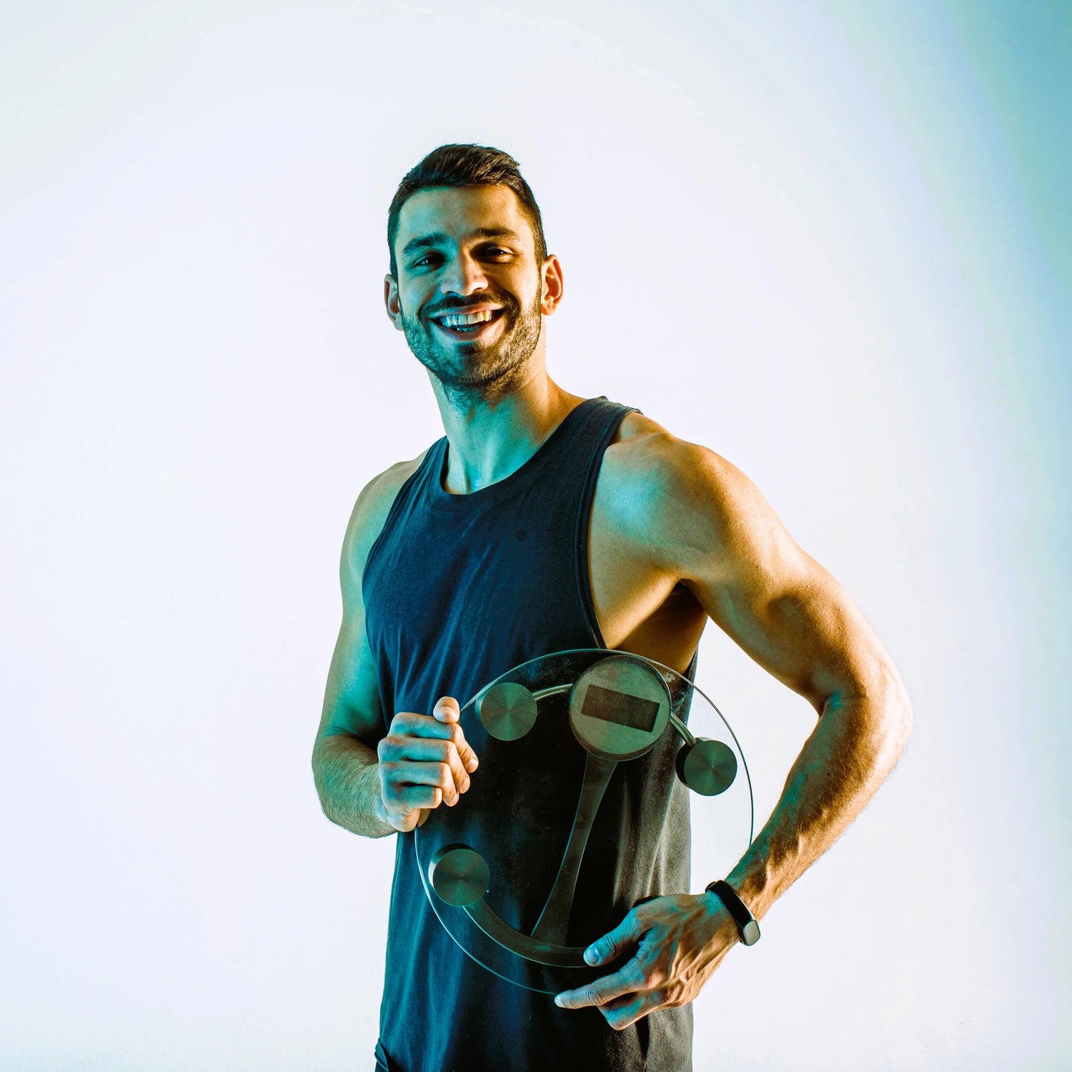 Sportieve jonge man in zwart hemd met weegschaal glimlacht op turquoise studio achtergrond